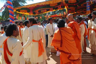A festival gathering beneath an open pavilion decorated with orange and yellow drapes, green-tipped leaf swags, orange triangular pennants and a tall balloon column of red, blue and orange balloons at left. Several men in cream and white kurta-style garments wear orange sashes tied at the waist, some with gold chains. Two monastics in orange robes stand at the centre-right, one bald with a draped orange shawl and the other with long dark hair and an orange garment, facing each other in conversation. Other men in white and orange cloth stand around them, and a few people are visible in the background among palm trees.