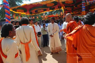 Outdoor festival scene under a large red and yellow canopy decorated with green, orange and yellow fabric swags and strings of small orange flags, with a palm tree and a balloon arch at the left edge. A crowd of young men and boys in white and cream kurtas, some with orange shoulder sashes, stands on a dirt ground. In the centre, a man in a cream kurta and white dhoti with a white shawl and a red-and-white bead necklace stands with his hands clasped. On the right, two bald monks in saffron-orange robes gesture toward him, one pointing with an open hand. On the left foreground, a man with curly hair wears a cream kurta with an orange sash. Several onlookers stand behind, some holding small items.
