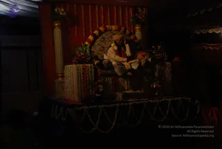 A dimly lit photograph of a seated figure on a zebra-striped throne-like seat set on a raised stage. The figure wears a cream kurta with a red shawl and a light turban, and sits with one hand raised near the face. The throne is framed by a red backdrop hung with garlands of marigolds and white flowers, flanked by white and gold pillars with flower arrangements, and the stage edge is draped with white jasmine garlands. The scene appears to be an evening celebration with a dark, sparse audience area.