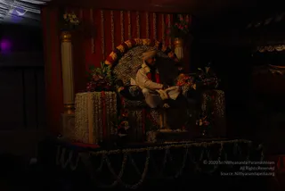 A dimly lit photograph of a seated man on a decorated throne at an event. He wears a cream or white kurta-style outfit, a cream turban, and a red-and-white garland draped over his shoulders. He sits on a zebra-striped chair framed by a curved arch of marigold and rose garlands, with red and orange flowers arranged beside the throne and on a stepped platform. A white jasmine garland hangs along the stage edge. Red drapery and a cream column stand behind him. The man's features are not clear enough to judge whether he is the a person; the appearance is uncertain, so the candidate status is unsure. One person is visible.