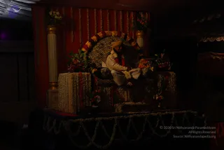 A dimly lit stage set with a zebra-striped throne draped in garlands of white and red flowers, with a red curtain backdrop strung with flower strands and two gold-and-white pillars topped with floral arrangements. A seated man in a cream-coloured silk kurta and white dhoti, wearing a pale turban and a red and white flower garland, rests with his hands on his lap while sitting on the throne. Floral garlands drape the stage edge and a small table of flowers stands at the side.