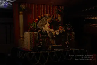 A dimly lit stage set for a ceremony. A seated figure in a white ceremonial garment and a cream turban sits on a zebra-striped throne with a floral garland draped across its back. The throne is framed by red fabric hung with marigold strings, white flower-covered pedestals, and vases of red and orange blooms. A flower-garlanded table in front holds a small lamp. The figure appears to be a single person seated in ceremonial dress, with the appearance of a person seated on the throne and no clear basis for identifying them.
