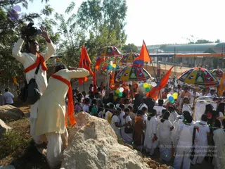 A festival procession seen from a rocky rise. In the foreground a man in white kurta with an orange shawl raises a video camera overhead, and beside him another man in white with an orange scarf gestures toward the crowd. A large crowd of people in white clothes moves along a dusty road lined with orange flags on poles and multicoloured umbrellas. Balloons in green, blue, white and yellow are tied to the procession, and strings of small lights run above the crowd. Among the people, a few wear orange garments. Sheds and a striped banner line the background under a bright sky.