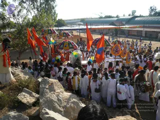 Outdoor procession at a wide dirt-ground ashram campus with metal-roofed sheds behind. Dozens of people in white clothing, many women and girls in white saris and men in white kurtas, walk past large rocks, following a decorated palanquin-like float. A drummer in a white outfit with a red sash plays a drum at right. Orange saffron flags on poles and multicoloured striped umbrellas rise above the crowd, and balloons in yellow, green and blue hang from the float. A cameraman in an orange scarf stands at the left edge on a rise. The float carries orange-robed figures near its top, and no one can be identified with confidence.