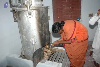 A photograph of a person in orange robes with a red border bending over a wood-fired stove. A stainless steel cylindrical vessel with pipes stands behind a small fire of split logs on a concrete hearth. The person in orange holds a brass lamp or vessel toward the flames, while a figure in white clothes is partly visible at the right edge. A red-brick wall and a water bottle sit in the background.