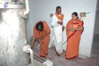 A photograph inside a bare, whitewashed utility room with exposed wiring. On the left, a large cylindrical stainless steel tank with pipes stands beside a woman in orange sari bending down to touch a pipe fitting; she wears a gold bangle and a long beaded necklace. In the centre, a man in a white dhoti and an orange shawl, with a tilak on his forehead, stands with his hands folded. On the right, a woman in an orange sari with red border holds a small silver vessel and looks down. All three are barefoot on a concrete floor. Three people are visible.