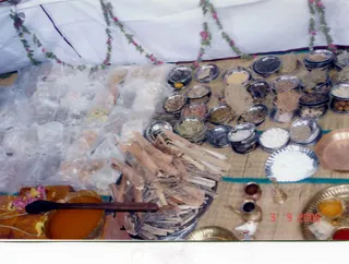 A photograph of an offering table set up under a white tent decorated with strings of pink and green flowers. Rows of small silver bowls hold assorted grains, powders, dried herbs and spices. Bundles of cut wooden sticks lie on a metal tray beside a bowl of thick orange liquid with a dark wooden ladle. A brass kalasha-style vessel, brass lamps, a small brass bell-shaped vessel, a bowl of white grains and a copper plate are arranged on the table. Plastic-wrapped packets of material sit at the left. No people are visible in the scene.
