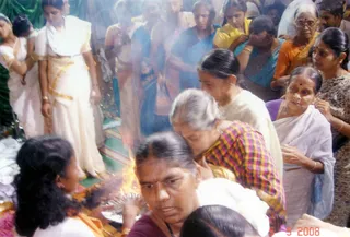 A crowded indoor ritual scene with a small fire burning at lower left, its flames rising near a metal vessel. Several women stand close together, some in white and cream saris with gold borders, others in blue, orange and yellow saris, and one woman in a pink-and-yellow checked blouse leans forward over the fire area. Another woman in a purple-edged white sari with gold bangles watches intently. A woman in front wears a white garment and dark maroon top and looks down toward the flames. Smoke drifts across the frame and a green cloth hangs in the background.