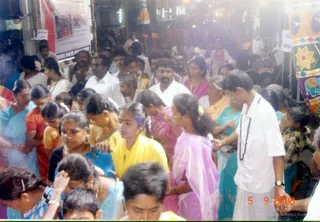A dense crowd of devotees fills a busy street or temple-side passage under bright overhead light. In the foreground a woman in a blue sari with gold bangles and a gold necklace bends forward, and a young woman in a yellow sari stands nearby. Beside them a young girl wears a pink-lilac sari, and a woman in a teal sari with a gold necklace stands at right. At far right a young man in a white shirt and white dhoti stands with a dark rudraksha-style bead mala across his chest. Men in white shirts and a striped plaid shirt appear among the crowd, and a red-and-white banner with pictures hangs on the left. A large colourful festival panel with floral and geometric designs decorates the right edge. The scene suggests a ritual procession or darshan line, with people pressed closely together.