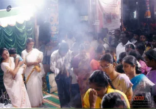 A dense crowd of devotees stands and kneels in a smoky, dimly lit temple hall during an evening puja. In the left foreground a woman in a white and gold-bordered saree holds a small child in blue. Beside her, another woman in a white saree with gold border stands with hands clasped. Center and right, women in pink, orange, blue and magenta sarees and men in white shirts stand close together, some with palms pressed in prayer. A girl in a yellow top in the lower foreground brings her hands to her face. Behind them are green draped cloth, a green-and-white canopy, and red and orange decorations and banners. Smoke or incense haze fills the air.
