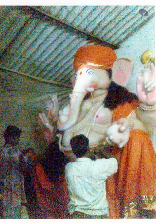 A large Ganesha murti dominates the frame, with white elephant head, orange crown-like headdress with gold band, and a raised hand holding a sweet or modak. Its body is draped in orange and gold cloth. In the foreground, a person in a white shirt and a few other figures in dark and red clothing stand before it, partly cropped. Ceiling slats are visible at upper left.