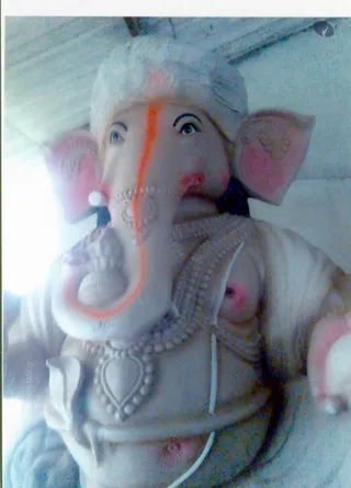 A close, upward-angled photograph of a large white clay Ganesha murti. The elephant-headed deity has an orange-red vertical line on its trunk and forehead, painted pink-tinged ears with ornamental patterns, and a beaded necklace with a heart-shaped pendant over its chest. Its trunk curls toward the lower left, and a small modak-like sweet is held near the trunk. No people are visible; the plain white background suggests an indoor setting with a ceiling above.