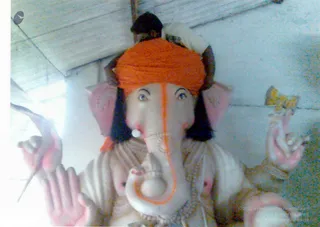 A photograph of a white Ganesha murti with an elephant head, orange tilak line down the trunk and forehead, and an orange turban-like cloth on the head. The deity has pink-tinted hands holding objects, a dark-haired headdress, and white beaded necklaces. A small figure with a dark head appears behind the murti under a plain ceiling. The image is taken close up, and the murti fills most of the frame.
