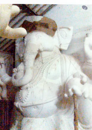 A close-up photograph of a white stone or marble sculpture of the elephant-headed deity Ganesha, seated with a trunk curling toward the lower left and a long ornate garland draped across the chest. Carved beaded necklaces and a decorated belt are visible, along with a raised hand holding a small object. Part of a second carved figure appears at the right edge. No living people are visible.