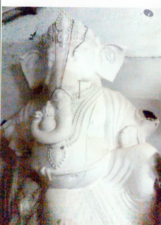 A pale, washed-out photograph of a white stone or plaster Ganesha murti with an elephant head, shown in profile. The figure wears a tall crown, a beaded necklace and a draped cloth, and its trunk curls toward the left. No people are visible.