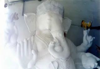A large white unpainted clay Ganesha murti, in an early stage of sculpting, shown in close-up. The elephant-headed deity has a broad trunk curling toward the centre, rounded ears, a cloth or plaster-wrapped crown, and multiple arms with the hands raised. Strings of beaded necklaces and a ribbed belly are modelled in relief. Behind it stands a wall with a yellow and orange object and a dark pole. No people are visible, and the image is a photograph of a sculpted deity figure.