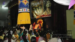 Photograph of an indoor event hall with a large crowd seated on the floor facing a stage. A large banner behind the stage shows a portrait labelled "Paramahamsa Nithyananda" beside gold-toned lettering. On the stage, a figure in a red robe stands near a decorated floral arch and a large portrait-like picture in an orange frame, while a woman in a red sari and a man in white robes stand beside it. In the foreground, many women in white, saffron and colourful saris sit with hands folded. A tall yellow and blue decorative hanging and a small brass lamp stand are visible on the sides.