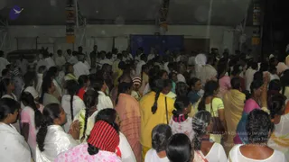 A dense crowd of devotees fills a dim tent-like hall, mostly women with long braids in white, cream, pink, yellow and green saris and salwar outfits, many with palms pressed together in prayer. Men in white shirts and dhotis stand among them toward the back. Vertical banners with decorative motifs hang on the side pillars, and a blue cloth backdrop hangs at the far end. No clearly identifiable a person figure is visible in this frame.