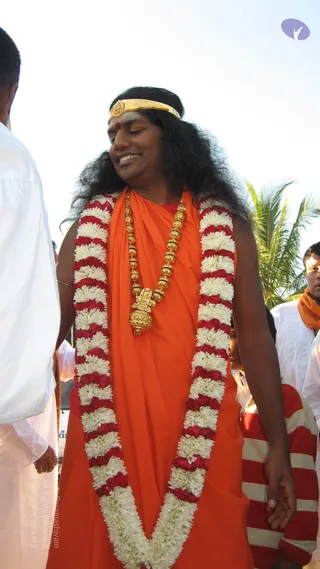 A smiling man with long dark swept-back hair stands outdoors in a saffron-orange robe, wearing a gold headband with a red bindi, a long gold bead necklace with an ornate gold pendant, and a thick garland of white and red flowers draped from his shoulders to his waist. Beside him a man in white clothing stands at the left edge, another man in white and orange appears at the right, and a palm tree is visible against a pale sky.
