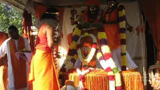 Outdoor photo of a puja at a temple-like dais draped in orange cloth. A figure in a red sleeveless top and orange dhoti stands at the center-left, arms raised toward the altar, with a long dark curly hairstyle and a gold headband. At left, a man in white dhoti and orange shawl holds a tray with flowers. Behind them stand two large statues in orange robes with a crown and trident-like staff, dressed with garlands of yellow, purple, red and white flowers. In front, a small figure in orange is adorned with flower garlands on an orange-fringed platform, with a gold-trimmed ornament at the right edge. Trees and sunlight appear in the background.