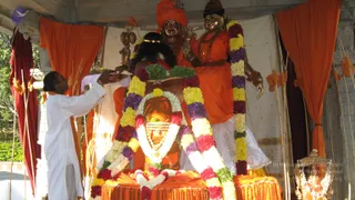 A priest in white dhoti and shirt stands at the left, holding a tray or cloth near a shrine adorned with multicoloured flower garlands in yellow, purple, red and white. Behind the garlands sit two large golden-brown statues in orange garments: a central figure wearing an orange turban-like crown and a standing or seated figure beside it with an orange robe and long garland. Orange and maroon cloth draping and bamboo poles decorate the open pavilion. In the front, an orange-draped altar holds a linga-like form with horizontal tripundra markings and a garland. The scene is a puja with abhishekam-style offerings at an outdoor shrine; the statues are depictions, not living people.