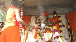 A photograph of a ritual at an outdoor pavilion draped with orange cloth. In the foreground a man with long dark hair and an orange garment holds a silver lamp with a handle, swinging it toward two figures. Behind him, two life-size statues in orange robes with garlands of white, yellow, purple and green flowers stand on a raised platform; one statue wears an orange turban and the other a dark crown, and both hold ceremonial objects. Smoke or incense rises between them. A second man with a beard, partly visible at the left edge, wears orange. The scene looks like an aarti or abhishekam performed before murtis.