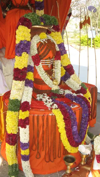A photograph of a seated figure. The figure wears a large rudraksha mala across the chest and waist, a gold-toned pendant on a chain, and a silver-toned ornament at the neck. A brass kalasha-style vessel and a brass lamp stand at the base, with white flowers and a conch-like shell offering in front. Dangling rudraksha strands hang from the orange seat, and tall bamboo poles and a potted green garden appear in the background. This is an idol, not a living person, so no physical people are counted.