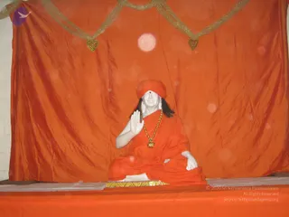 A white marble statue seated in lotus-style posture on a golden-edged plinth before an orange draped backdrop with gold tinsel swags and heart-shaped gold ornaments. The figure wears an orange saffron wrap, an orange head cloth with a red bindi on the forehead, a long dark hair, and a gold necklace with a pendant. Its right hand is raised in abhaya-style blessing while the left rests on the lap. An orange draped table front and a faint circular light glow appear in the scene. The statue is a depiction, not a living person.