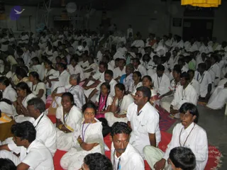 A large seated gathering of devotees in an indoor hall, mostly dressed in white, with many wearing rudraksha malas around their necks. They sit on red mats and rugs across the floor, facing forward toward an unseen focal point. Women in white saris with coloured borders and a man in a white shirt with a mala are in the foreground; rows of people fill the background. Yellow fabric hangs from the ceiling at upper right and a few people stand near the back. Dim lighting and overhead rigging are visible.