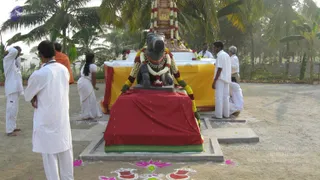 Outdoor photo of a black stone Nandi bull statue seated on a square plinth draped in red cloth with a green-and-gold border, decorated with white and yellow flower garlands and a red-and-white necklace. Behind it stands a tall structure with a white base wrapped in yellow cloth, topped by a garland-wrapped golden shrine element, set among palm and banana trees. Several men in white dhotis and shirts stand around, one in an orange garment at left, and a woman in a white sari stands at left center. Colourful rangoli kolam designs in pink, green, and red decorate the sandy ground at the bottom.