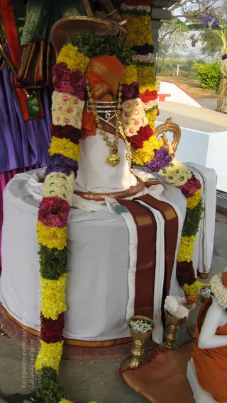 A photograph of a statue or idol of Swamiji, the Nithyananda Paramashivam, seated on a white draped pedestal, adorned with long garlands of yellow, red, pink and purple flowers with green leaves. The figure wears an orange and gold turban, a gold ornamental necklace with a pendant, and a white dhoti with a maroon and gold border. Brass oil lamps with offerings sit at the base, and a white-clad figure in orange kneels nearby. Behind is a green and purple cloth backdrop and a bright outdoor setting with trees. This is a depiction in statue form, not a living person.
