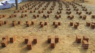 A photograph of many stacked terracotta-red bricks arranged in rows across a sandy, open ground. The bricks sit in neat clusters of two to four, casting shadows on the pale sand. At the upper left, a partly visible person in dark clothing sits behind a stack of white sheeting and a palm tree trunk. No ritual activity, deity or a person depiction is visible.