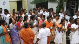 A crowd of devotees stands outside a white building with an open green door, holding small metal pots and vessels aloft. Many women wear saris and salwar kameez in orange, white, red, blue and teal, with orange shawls draped over shoulders, while men wear white shirts and dhotis. A banana leaf hangs over the doorway beside a black board listing names of devotees who built Ananda Sabha, with the year 2005 at the bottom. A young boy in a white shirt balances a kalasha-style pot on his head. Some devotees wear lanyards and rudraksha-style beaded necklaces. No single clearly identifiable a person figure is visible among the people shown.