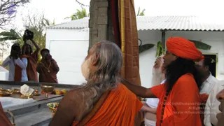 Outdoor ritual photograph at a stone-pillared shrine with a white-washed building and corrugated roof behind. In the centre, an elderly bearded man with long grey hair and an orange-saffron garment stands with his back to the camera, facing a draped gold-bordered cloth. To the right, a younger man in an orange turban and orange top with a rudraksha mala stands close beside him, one hand extended toward the shrine. On a stone platform in the foreground sit brass bowls and lamps, a white conch shell, a bowl of fruit and flower offerings. On the left, several photographers in orange clothing hold up cameras, and a white-clad man and a bearded man in white stand at the right edge.