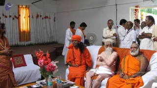 Indoor photo of a white-walled hall with a red-and-yellow striped curtain and a row of ceremonial staffs with tassels on the left wall. In the centre, a man with long dark hair, a saffron-orange turban and saffron robes with a rudraksha mala sits on a white sofa beside a white-bearded elder in a white loincloth and an elderly saffron-robed man. Standing men in white dhotis and orange shawls, and others in white shirts, gather behind them. A flower vase with pink blooms and water bottles sit on a low table with books. Swamiji (probable) is seated centrally, identified by long swept hair, a saffron turban, and rudraksha mala with saffron robes.