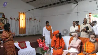 Photograph inside a plain white-walled hall with a corrugated roof, an orange-and-yellow striped curtain, and a row of long wooden staffs with orange tassels on the wall. At left, a framed portrait of a bearded figure in orange with a garland hangs beside a small altar. A man in an orange robe and saffron shawl stands at left, gesturing with his hands. Seated centre on a white sofa is a figure in an orange robe with a large orange turban, long dark hair and a rudraksha mala, looking to the side. Beside him sits an elderly white-bearded man with a bare chest and white dhoti, holding a cane. At right, an elderly man in orange sits on the sofa. Standing men in white shirts, a white kurta and orange shawls fill the background, and a pedestal fan and a bouquet of pink flowers sit in front of the sofa.
