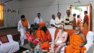 Indoor photo in a plain white-walled hall with orange curtains and a row of wooden rods hung on the left wall. A woman in an orange sari with an orange headwrap and a long rudraksha mala sits centre-left on a white sofa with her hands folded, a pink flower arrangement and water bottles on the low table in front. Beside her, an elderly bare-chested man with long white beard and a white loincloth sits on an orange-checked cushioned sofa with a cane, and an older man in an orange robe with white beard sits at the right edge. Behind them, several men in shirts and trousers stand watching, and a woman in orange and a man in an orange robe stand in the open doorway on the right. A man in white kurta with an orange shawl walks in from the left. No single figure is clearly a person by appearance, so identity is left unsure.