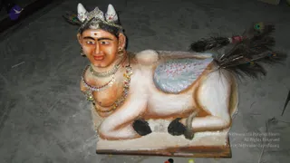 A photograph of a sculpted figure resembling a human head on a recumbent animal body, painted in pale cream and beige tones on a grey concrete floor. The head wears a crown with two white horn-like projections and jewelled ornaments, a red and white tilak on the forehead, and gold earrings. Multiple layered necklaces and beaded garlands hang over the chest. The body has a bluish-grey wing-like panel on the back and a tail ending in dark peacock feathers with eye patterns. Black hoof-like markings appear on the legs. Small props and a loose feather lie nearby. This appears to be a statue or idol rather than a living person, so no physical people are present.