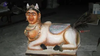 A photograph of a large sculpted figure with a human face and bull-like body, lying on a wooden platform in a dim storage area. The figure has white and tan painted skin, white horns with a gold crown, a tilak on the forehead, and wears layered gold and red beaded necklaces and a jewelled chest ornament. A decorated blue-and-pink saddle cloth covers its back, with a peacock feather tail. Behind it are cardboard boxes, sacks and a wooden table.
