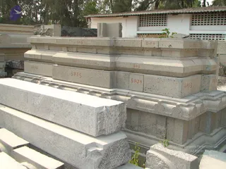 Photograph of stacked pale grey granite blocks and moulded temple-style stone components laid out in an outdoor yard. Several blocks bear red painted codes such as S.P.J.3, S.P.J.4, S.P.J.5, S.P.4 and S.P.6, marking them for assembly. Long slabs lie in the foreground, and a white building with ventilation grilles and trees stands behind. No people are visible.