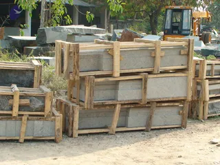 A photograph of a stone yard with stacks of cut grey granite blocks strapped into rough wooden crates and pallets on bare dirt ground. Large uncut grey boulders lie behind the crates, with a yellow wheel loader with a cab at the upper right and a corrugated-roof shed and green trees in the background. No people are visible.