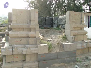 A photograph of stone construction at a temple-building site. Pale granite blocks carved with pillars, mouldings and pilasters are stacked in tiers, with orange alphanumeric markings painted on each block such as G1, G2, G3 and G.K.2. Loose dirt, scattered wooden planks and debris lie in the foreground, with dark rough-cut boulders piled behind and tall trees rising above. A white building with windows stands at the right edge. No people are visible.