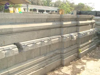 A photograph of carved grey granite temple base mouldings stacked outdoors in a yard, with stepped horizontal courses and ornate moulded ledges with projecting cornice details. A tarpaulin canopy, a plain white building, palm and tree foliage and dry leaves on the ground surround the stones. No people are visible in the scene.