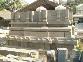 A photograph of granite temple construction pieces stacked outdoors in a yard beneath a thatched shelter roof, with palm trees behind. The stacked stones are carved architectural elements, including moulded cornice blocks, pilasters and small domed finials, many labelled in red paint with codes such as Gr.13, Gr.12, Gr.11, Gr.K.9, Gr.P.J.9 and Gr.P.J.10. Loose cut stone blocks and rubble lie in the foreground, and a white stone pillar and carved sections sit at the right edge. No people are visible.