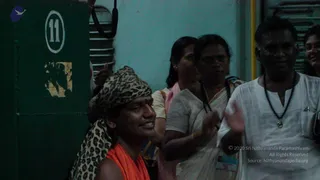 A photograph taken in a dim indoor corridor beside a dark green door marked with a white circled number 11. In the foreground a man wearing an orange saffron garment and a leopard-print cloth wrapped around his head smiles toward the camera. Behind him a woman in a pink top and a woman in a white sari with a dark beaded necklace stand near a man in a white kurta wearing a dark bead necklace with a pendant and an ID badge. A woman in glasses appears at the right edge. The foreground man's identity is uncertain from appearance alone, so he is described by role only.