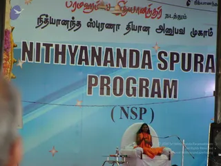 A photograph of a seated speaker on a white armchair-style throne on a stage, dressed in saffron-orange robes with long dark hair, holding his hands together and speaking into a microphone on a stand. A blue stage backdrop reads NITHYANANDA SPURANA PROGRAM (NSP), with a Tamil banner above it. A small table with a water bottle sits to the left, and a speaker cabinet is at the right edge. A blurred person's shoulder appears in the lower left foreground.