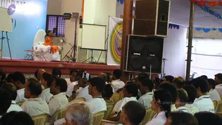 A photograph of a seated audience facing a raised stage at an indoor hall. On the stage, a man with long dark hair and an orange-saffron garment sits on a white throne-like chair with a small table and water bottles beside him. Many attendees in white shirts sit in plastic chairs, several wearing headphones for translation. A large black speaker cabinet stands at right, and a white banner with a colourful concentric circular design hangs beside the stage. Blue and orange bunting decorates the ceiling, and a projection screen stands behind the stage.