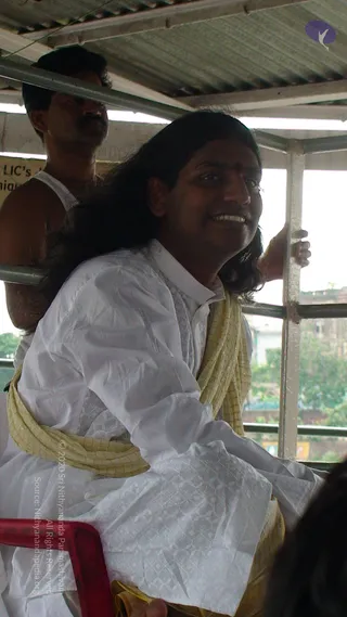 A photograph of a long-haired man in a white kurta with a pale gold shawl draped over his shoulders and wrapped around his waist, seated on a red plastic chair under a corrugated metal roof. He smiles and looks back over his shoulder toward the camera. A second man in a white vest stands behind him, one hand gripping a metal rail. Metal frame posts and a green city background are visible through the open side. Swamiji (probable) is shown in person, identified by long swept-back hair, a red forehead dot, and gold-bordered shawl cues; the seated man is the candidate.