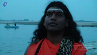 A photograph of a long-haired man in an orange saffron robe with a leopard-print shawl draped over one shoulder, standing on a riverbank at dusk. He has a white horizontal tilak and a red dot on his forehead and looks upward. Behind him a small boat with a few people sits on calm, blue-grey river water, and a flat sandy shore stretches along the far bank.