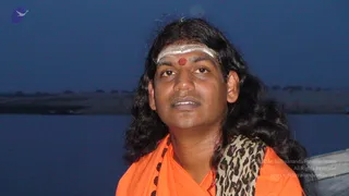 Probable Swamiji, a man with long, swept-back dark hair, wearing saffron-orange robes and a leopard-print shawl over one shoulder, stands before a river at dusk. He has white vertical tilaka marks on his forehead with a red dot at the centre, and a rudraksha mala is visible at his chest. The water and a flat far shoreline fill the background under a deep blue evening sky.